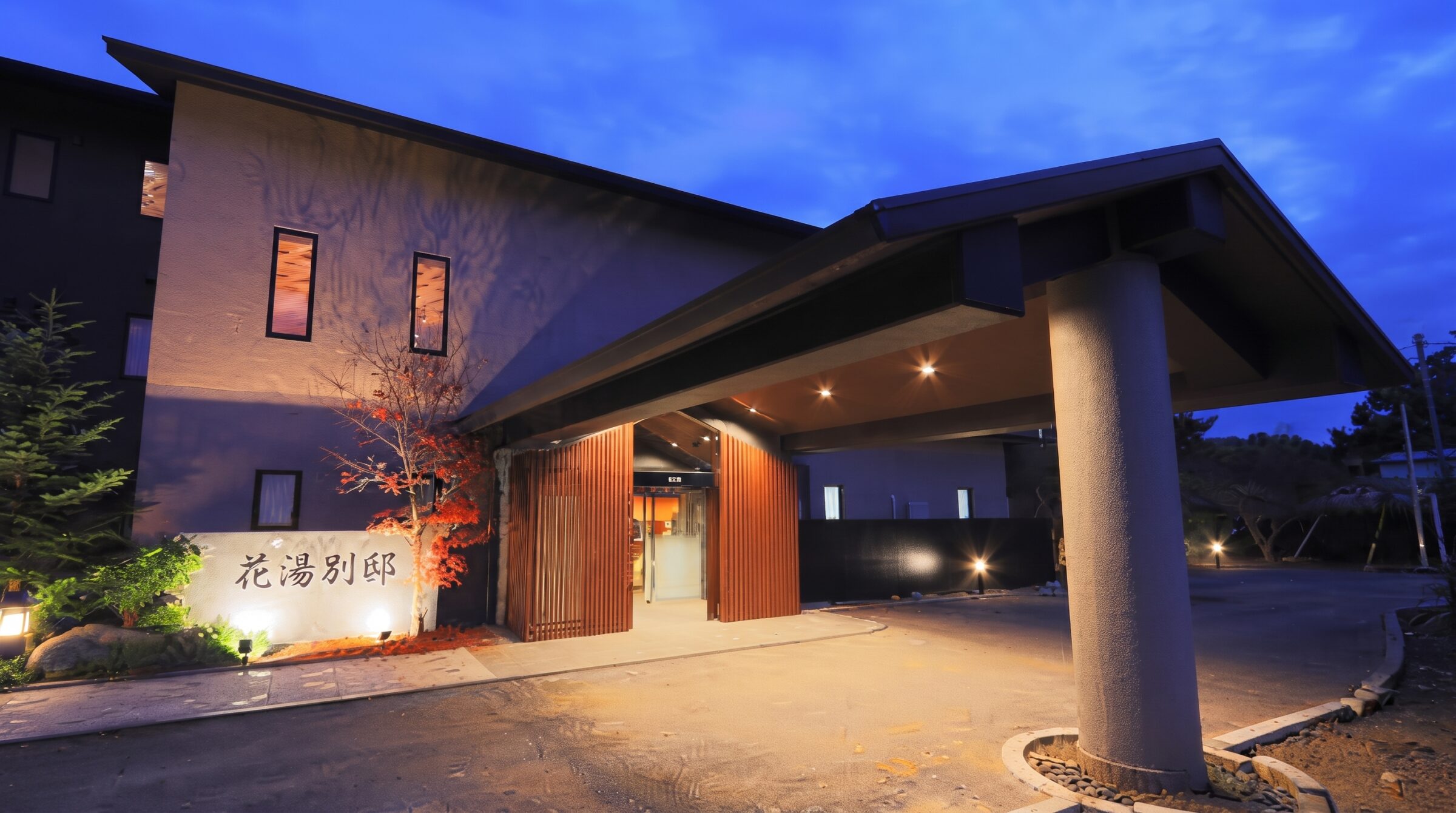 夕暮れ時に温かい照明が灯り、「花湯別邸」の看板が掲げられたモダンで洗練された宿のエントランスと外観の様子